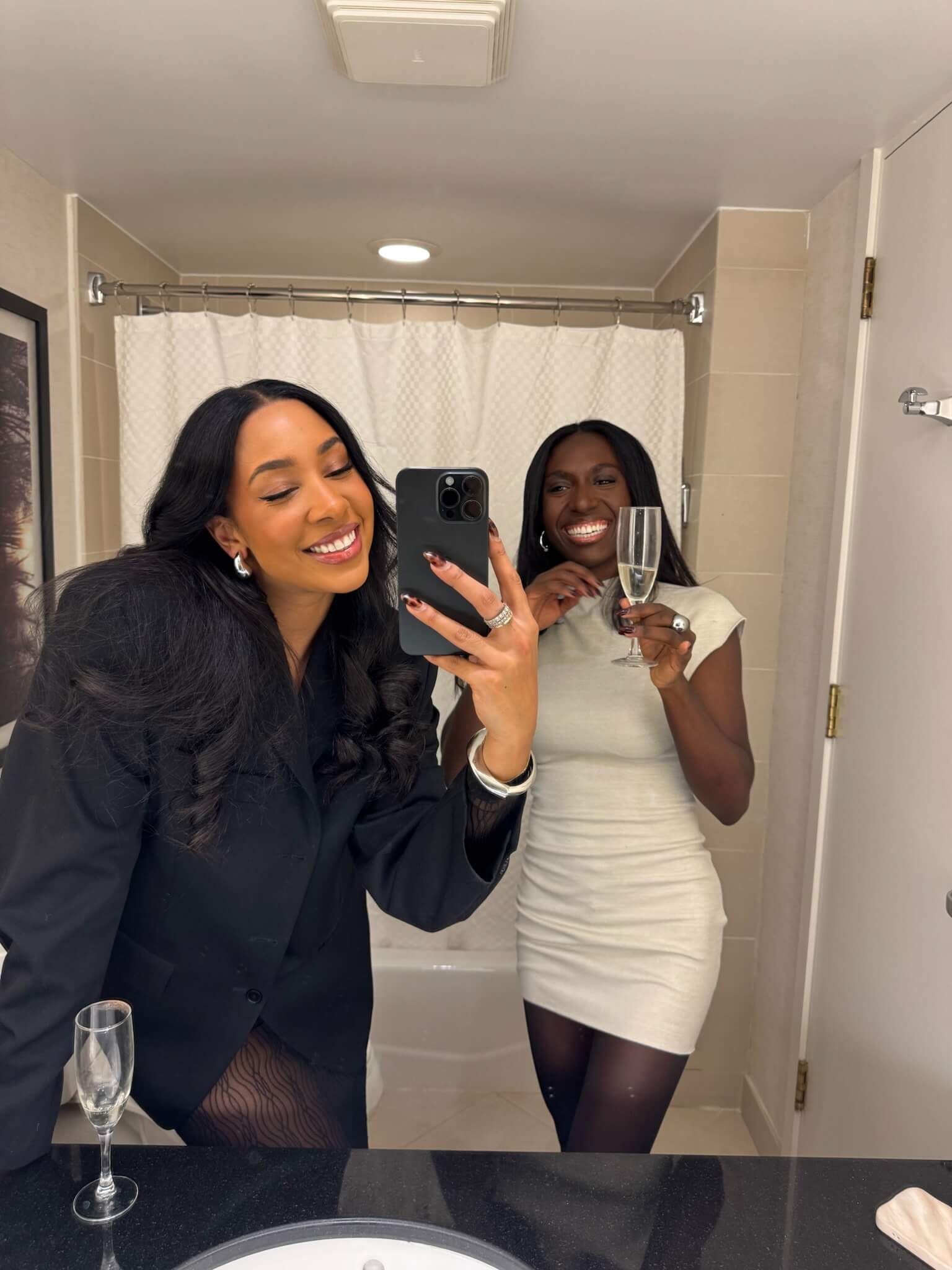 Two black women with hair extensions taking a mirror selfie in a bathroom with drinks.