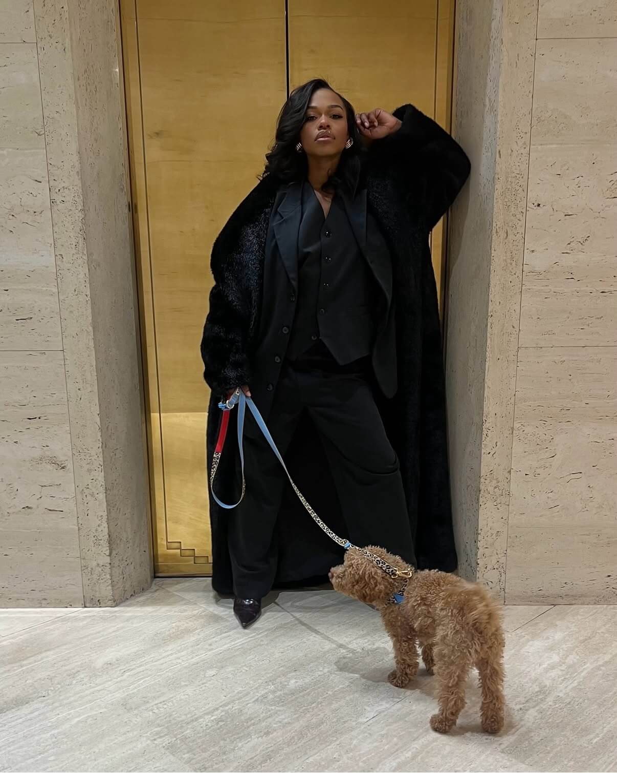 Black woman with short curled hair extensions wearing black coat with a dog on a leash against a stone wall.