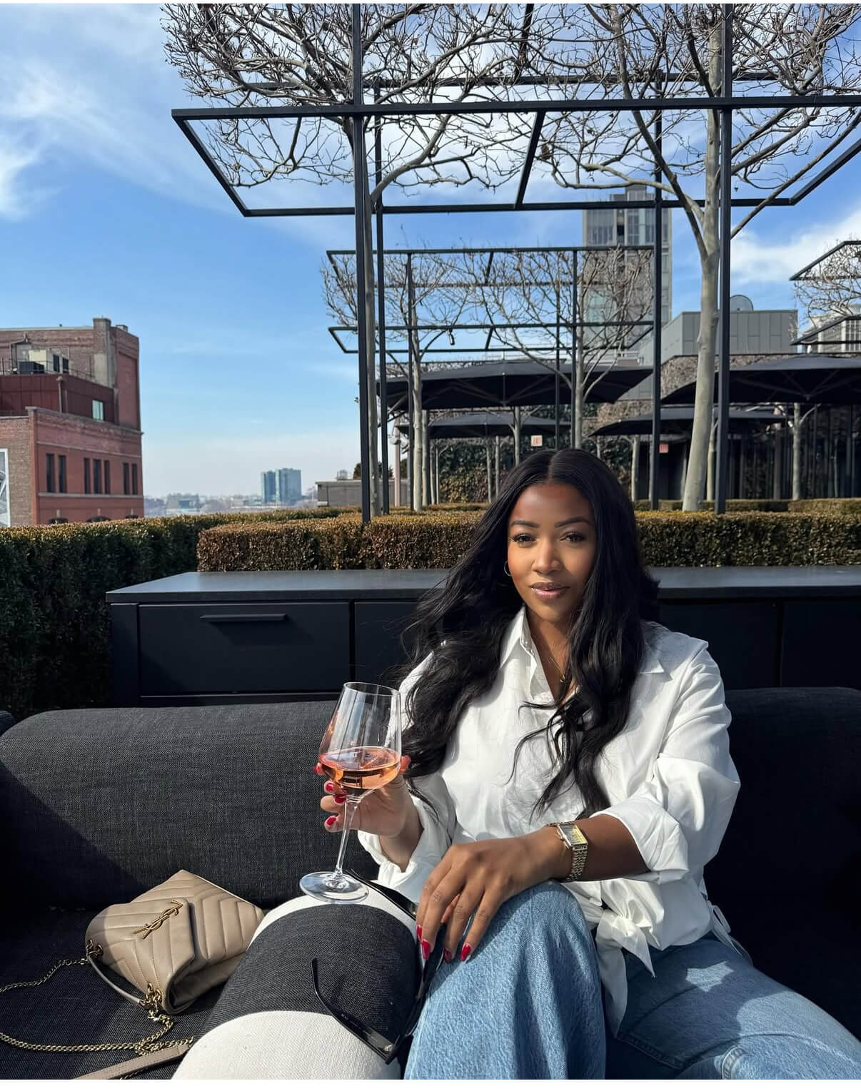 Black woman wearing SOWN body wave hair extensions bundles sitting on a rooftop with a glass of wine, surrounded by urban city scenery.