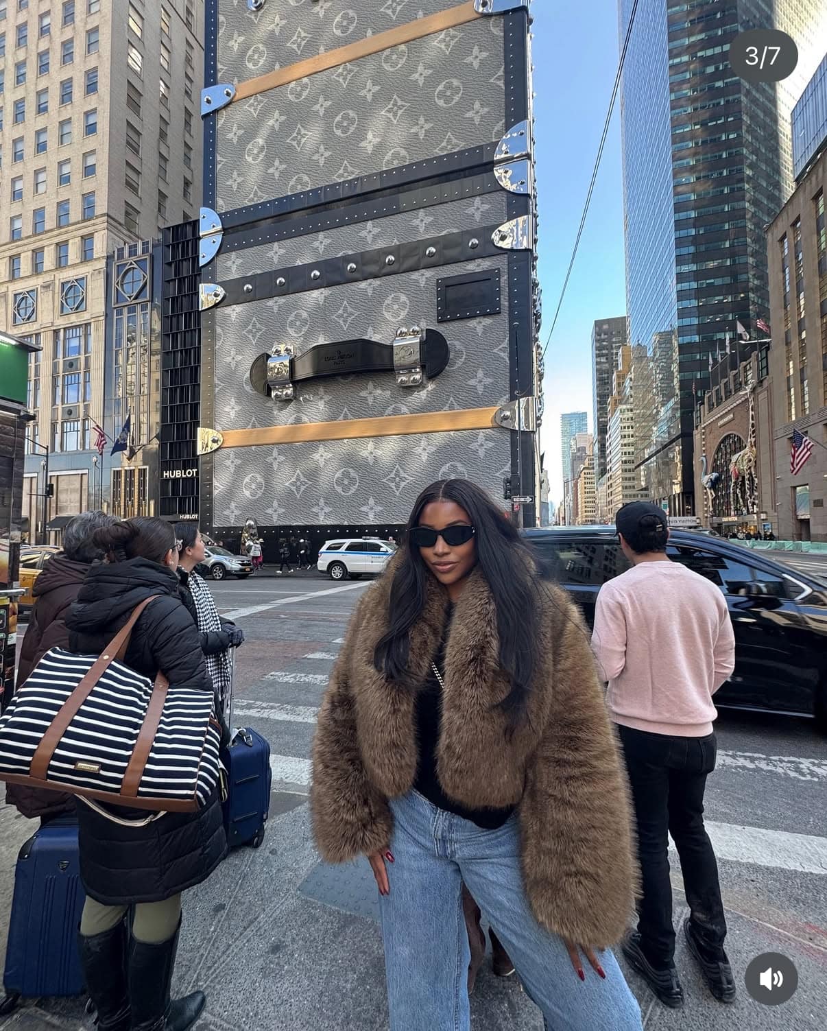 Black woman wearing SOWN hair extension dressed in fur coat standing in front of a large Louis Vuitton suitcase on a city street.